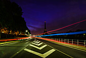 A view along a city road at night, tall buildings and light trails, the Eiffel tower in the distance.