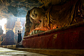 Ein Höhlentempel, eine große liegende Buddha-Statue, brennende Kerzen und ein betender Mönch, Myanmar, Asien