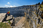 Two people hiking stand at a cliff and look at the Creux du Van rondel, Swiss Jura, Neuchâtel, Switzerland