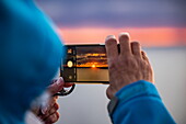 Frau macht Sonnenaufgangsfoto mit Smartphone in den Stockholmer Schären, Stockholmer Schären, in der Nähe von Stockholm, Schweden, Europa