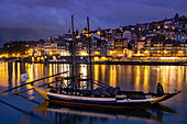 Traditionelles Portwein-Transportboot auf dem Fluss Douro mit Blick auf die Altstadt von Ribeira und das historische Zentrum in der Abenddämmerung, Vila Nova de Gaia, Porto, Portugal, Europa