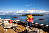 Ein Paar umarmt sich an der Reeling auf dem Deck von Expeditionskreuzfahrtschiff World Voyager (nicko cruises) mit Küste dahinter, Mariehamn, Alandinseln, Europa