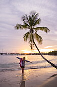 Woman stretches her arms and &#39;flies&#39; along L&#39;Anse Aux Epines beach with slate coconut palm at sunset, near Saint George&#39;s, Saint George, Grenada, Caribbean