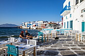 Woman and man relaxing on the sea wall while the cat stalks past empty chairs and tables in front of the seafront tavernas in &#39;Little Venice'39;, Mykonos, South Aegean, Greece, Europe