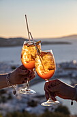 Hands of two women toasting with Aperol Spritz cocktails at 180º Sunset Bar on the hill overlooking the town and islands, Mykonos, South Aegean, Greece, Europe