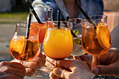 Close up of hands holding and toasting Aperol Spritz cocktail glasses at Stadtstrand bar, Wertheim, Spessart Mainland, Franconia, Baden-Wuerttemberg, Germany, Europe
