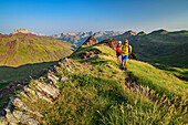 Man and woman hiking across ridges of grass, Castillo d&#39; Acher in background, Valle de Hecho, Huesca, Pyrenees, Aragon, Spain