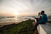 Touristen mit Gitarre am Abend an der Atlantikküste blicken aufs Meer, Algarve, Portugal