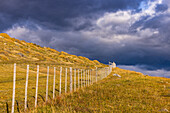 Perspective with a fence in the grassy landscape on a hillside in Patagonia in front of dramatic clouds, Chile, South America