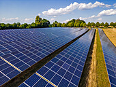 A ground mounted system of many solar panels on an open space with trees and clouds in the background, Hesse, Germany