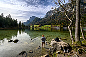 View of Hintersee, young man standing in the lake, shoes standing on the shore, wall path, nature trail on the stream/river Ramsauer Ache, hiking in the magic forest on Hintersee in the mountaineering village of Ramsau. Ramsau near Berchtesgaden, at the Watzmann and Königssee, Berchtesgaden National Park, Berchtesgaden Alps, Upper Bavaria, Bavaria, Germany