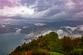 Luftaufnahme über die Stadt und den Luganersee im Tal mit Berglandschaft mit Sturmwolken in Lugano, Tessin, Schweiz.