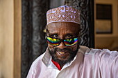 Lächelnder Mann mit muslimischem Taqiyah-Hut und bunt reflektierender Sonnenbrille, Lamu, Insel Lamu, Kenia, Afrika