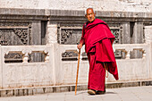 Tibetischer Mönch in rotem Gewand mit Stock geht an einer verzierten Mauer des Kloster Kloster Kumbum Champa Ling entlanf, Xining, China
