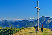 Mann und Frau beim Wandern stehen am Gipfelkreuz des Kratzer, Hohe Tauern im Hintergrund, Hirschberg, Bayerische Alpen, Oberbayern, Bayern, Deutschland