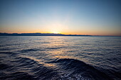 Blick auf Wellen und Meer mit Küstenlinie in der Ferne bei Sonnenuntergang, auf See, in der Nähe von Peloponnes,  Ägäis, Griechenland, Europa