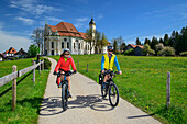 Mann und Frau fahren auf dem Bodensee-Königssee-Radweg mit Wieskirche im Hintergrund, Wies, UNESCO Welterbe Wieskirche, Oberbayern, Bayern, Deutschland