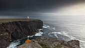  High cliffs by the sea with Stennes lighthouse at dusk. Rock with red lichen in the foreground. 