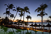 Strohschirme mit Kokospalmen am Strand bei Sonnenuntergang, Royal Palms Beachcomber Resorts, Grand Baie, Rivière du Rempart, Insel Mauritius, Indischer Ozean