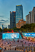 People walk in the Huacheng Square surrounded by modern high-rise buildings illuminated at dusk. Guangzhou, Guangdong Province, China.