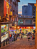  Menschen gehen in der beleuchteten Rua de S. Paulo (Dasanba) in der Abenddämmerung. Macau, China. 