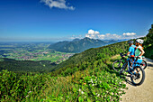  Man and woman mountain biking taking a break and looking at the Alpine foothills, Hochplatte, Chiemgau Alps, Upper Bavaria, Bavaria, Germany 