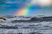 A vibrant rainbow arches over a turbulent ocean with crashing waves under a cloudy sky.