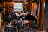 A vintage cobbler's workshop with wooden walls, filled with old tools and machinery for shoe repair. The room is cluttered with various equipment, a workbench, and a small window.