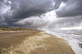 A lone person walks on a deserted beach under a dramatic cloudy sky, with waves crashing on the shore.