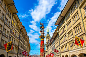 Beautiful Old City Street with Old Building and Flags and Clock Tower and a Statue in a Sunny Summer Day in City of Bern, Canton Bern, Switzerland.