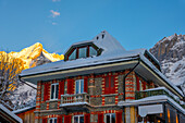 Historisches buntes Haus und Bergkette an einem verschneiten Wintertag bei Sonnenuntergang, Grindelwald, Kanton Bern, Schweiz.
