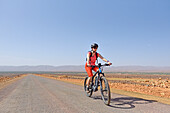 Young woman mountain biking with electric assistance on a straight road through the reg, from Agdz to Zagora, Draa River valley, Province of Zagora, Region Draa-Tafilalet, Morocco, North West Africa