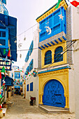  Alley with blue ornate wooden doors, Arabic balconies and windows, and colorful flags in the Medina of Hammamet, Tunisia 