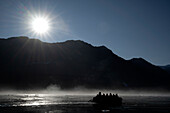  Backlit scene of the sun setting over dark mountains reflecting on fog and passengers in a Zodiac from an expedition cruise ship in the Vikingebugt, Arctic Greenland, Europe 