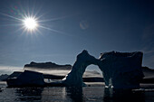  Backlit scene with the sun shining on the landscape and the passengers of an expedition cruise ship in a Zodiac rubber dinghy, seen through the bow of a large iceberg in the Vikingebugt, Arctic Greenland, Europe 