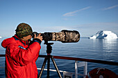  A photographer uses a tripod and a long lens with camouflage cover to photograph icebergs from the outside deck of an expedition cruise ship en route to Vikingebugt, East Greenland, Greenland Europe 