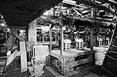 A black and white photograph of an industrial setting, showcasing machinery and equipment in a factory. The image captures the intricate details of pipes, metal structures, and a busy production environment.