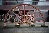 A large, rusted industrial wheel structure, resembling a giant fan or water wheel, set against a backdrop of brick buildings. The scene conveys a sense of history and industrial heritage.