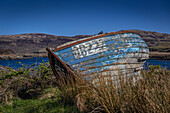 A small, dilapidated blue wooden fishing boat sits on dry land. Mountains and sea are in the background. 