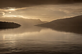 Goldene Morgenstimmung mit einer Wolkenlücke am Ufer von See Loch Katrine. Die Wolken und Berge spiegeln sich im Wasser, Schottland, Großbritannien