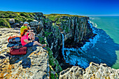  Man and woman hiking sitting on cliff overlooking Mfihlelo River waterfall, Wild Coast Trail, Wild Coast, Eastern Cape, South Africa 