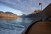  Man stands on the aft deck of the expedition cruise ship World Voyager (Atlas Ocean Voyages) with mountains at sunset, near Evighedsfjord (Eternity Fjord), Queqatta, Greenland, Europe 