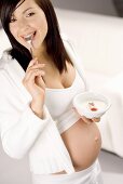 Schwangere Frau hält Schale Naturjoghurt und Löffel im Mund