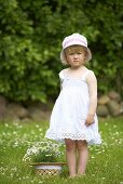 A little girl in a meadow standing next to a straw hat filled with daises