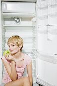 Junge Frau isst einen Apfel vor leerem Kühlschrank