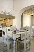 A dining table and chairs made of carved, white painted wood with a view through an archway into a living room
