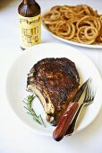 Rib Eye Steak on a White Plate; Rosemary Sprig; Knife and Fork; Onion Rings