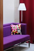 A view of a purple sofa with a colourful cushion and a floor lamp behind