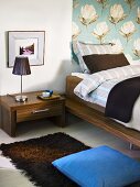 Corner of a bedroom with night stand made of dark wood and a wall hanging with a floral design in front of a bed