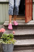 A child wearing pink shoes standing by a front door with a potted plant at the bottom of a three stone steps